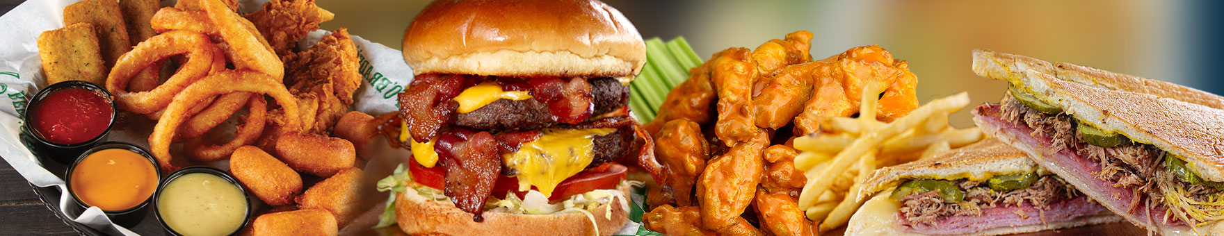 A juicy bacon hamburger is flanked on either side by a basket of wings, a cuban sandwich, and an assortment of starter items including onion rings and sauces